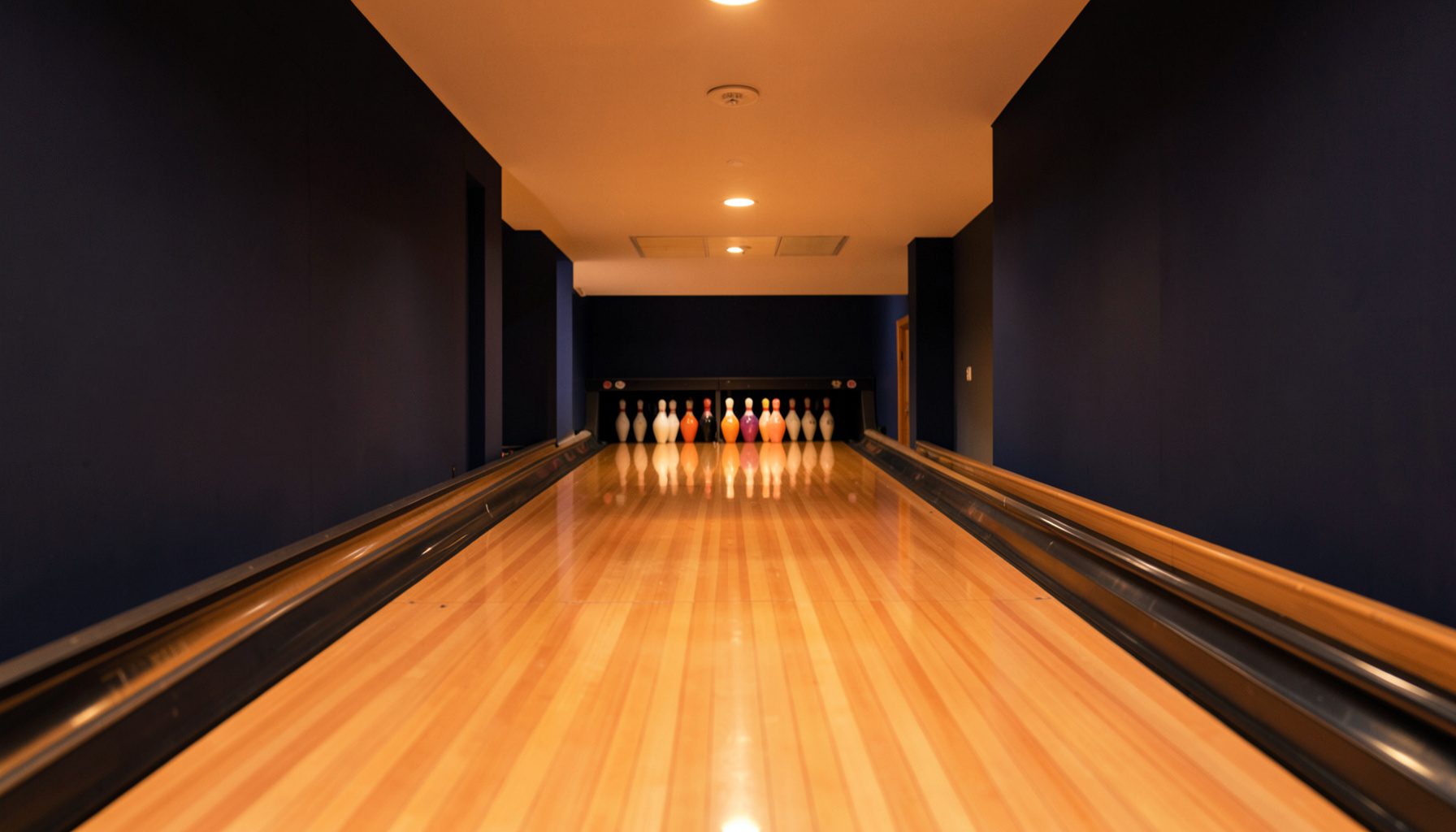A polished wooden bowling lane stretching toward a rack of pins, warm amber lighting reflecting off the lane surface, deep navy accents on the walls